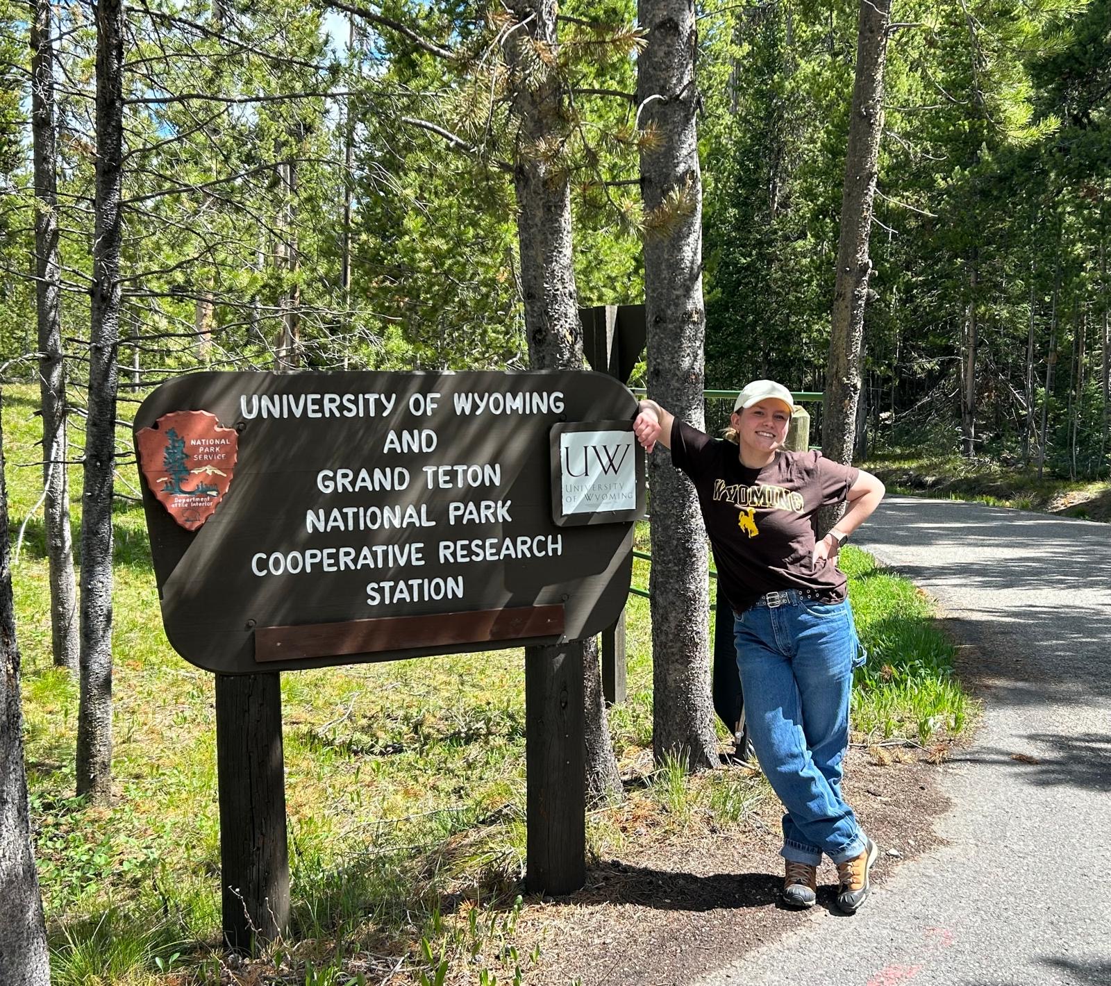 A woman with a brown shirt and jeans leans on a large wooden sign reading "University of Wyoming and Grand Teton National Park Cooperative Research Station"